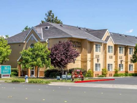The image shows a yellow two-story hotel building with a sign that reads "Quality Inn & Suites" surrounded by trees and a clear sky.