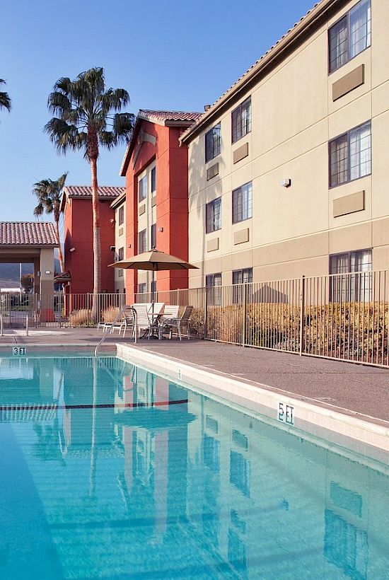 An outdoor pool surrounded by a fence, with a building and palm trees in the background under a clear sky.