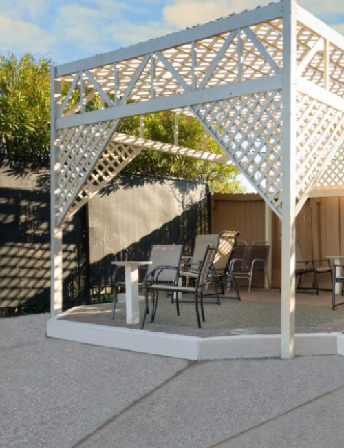 A pergola with seating on a patio, surrounded by a fence and plants, with a grill on the right side.