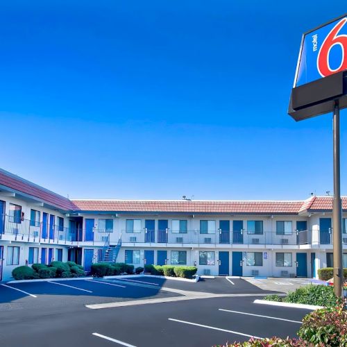 The image shows a motel exterior with a sign displaying the number 6, surrounded by a parking lot and clear blue sky.