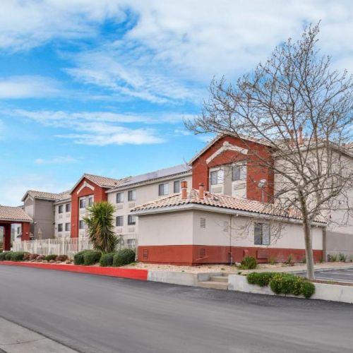 The image shows a two-story hotel building with red and beige exterior, surrounded by landscaping and a clear blue sky.