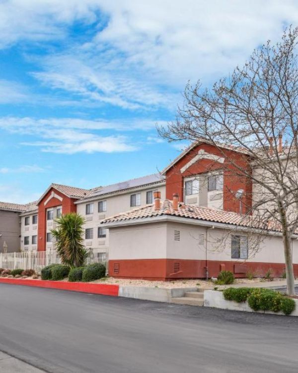 The image shows a two-story hotel building with red and beige exterior, surrounded by landscaping and a clear blue sky.