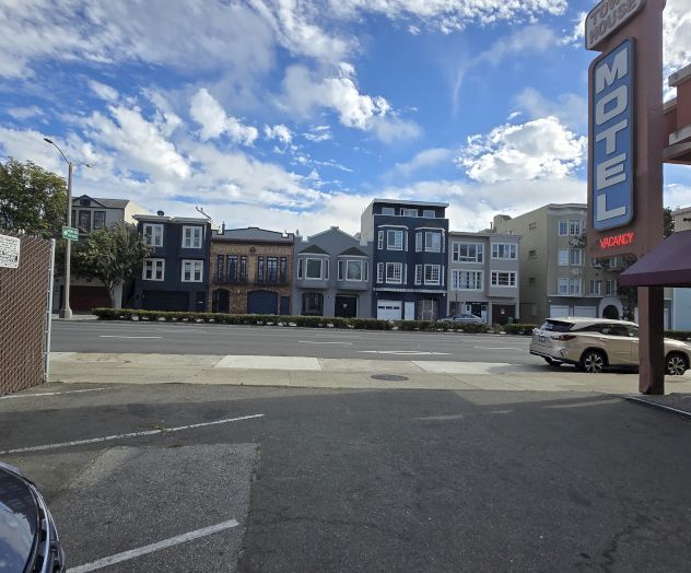 A motel parking lot with a vacant space and several buildings in the background under a partly cloudy sky.
