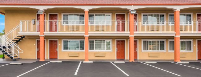 The image shows a two-story motel with external corridors, red doors, and an empty parking lot under a blue sky.