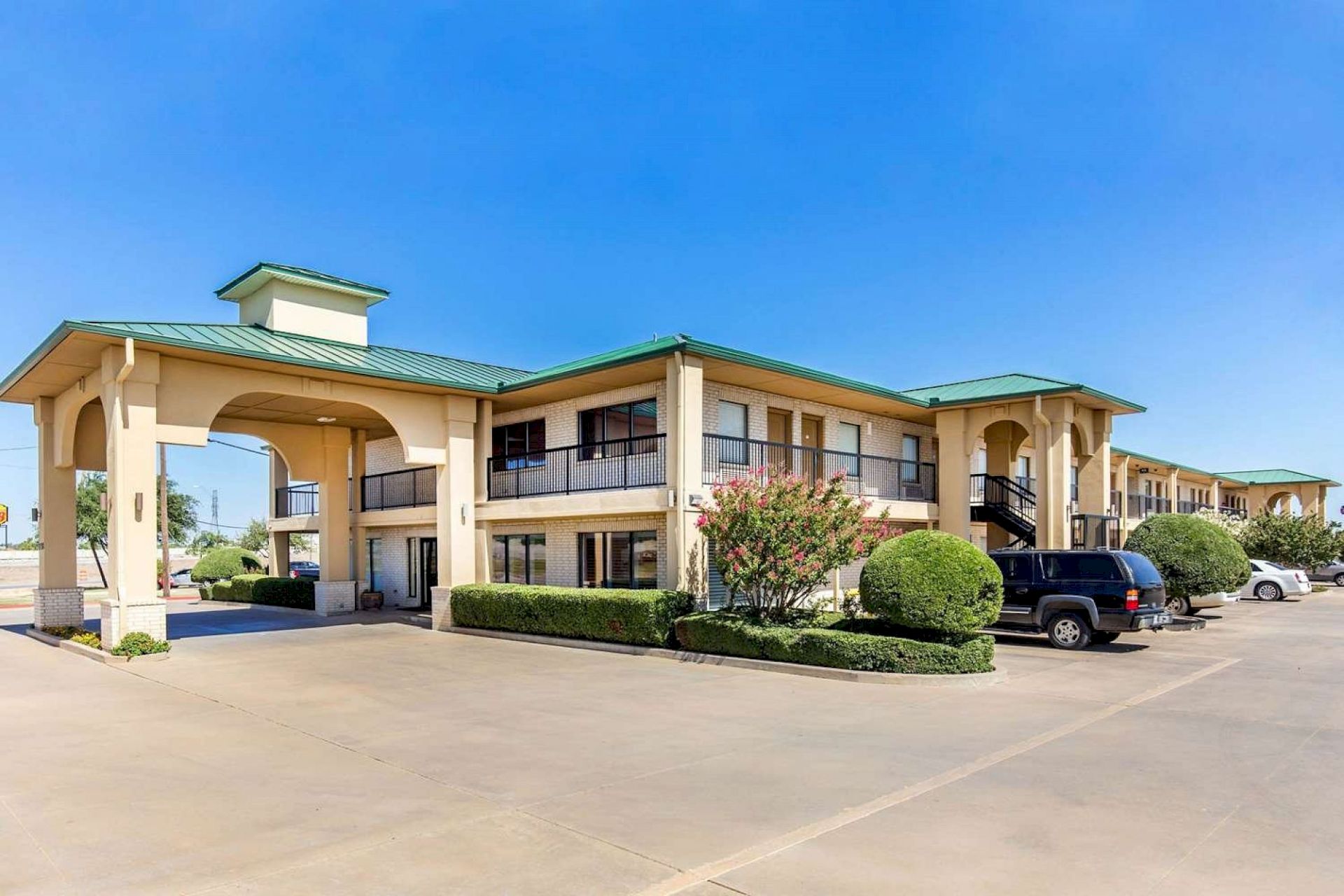 The image shows a two-story hotel building with a green roof, surrounded by a parking area and landscaped shrubs.