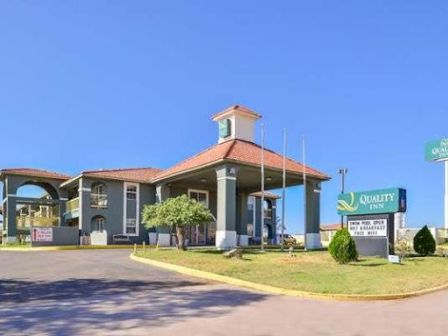 The image shows a Quality Inn hotel with a prominent sign, a paved entrance, and landscaped greenery under a clear blue sky.