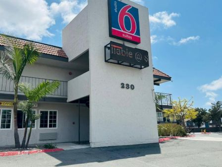 The image shows the exterior of a Motel 6 building with the sign and part of the parking area visible under a clear blue sky.