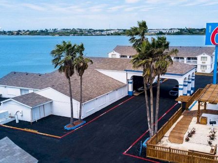 The image shows a Motel 6 near a body of water with palm trees and a pergola-covered area, captured on a sunny day.
