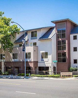 The image shows a modern three-story apartment building with large windows, a sloped metal roof, and a tree-lined street.