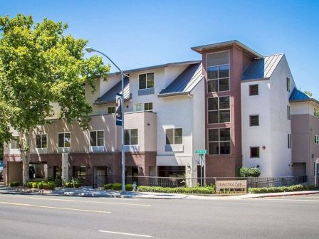 The image shows a modern three-story apartment building with large windows, a sloped metal roof, and a tree-lined street.