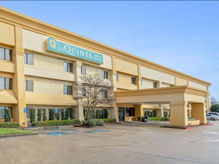 The image shows a La Quinta hotel building with a parking area, trees, and a clear, sunny sky above.