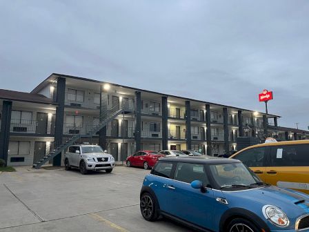 A three-story motel with external staircases and several parked cars. A bright red sign is visible on top of the building.