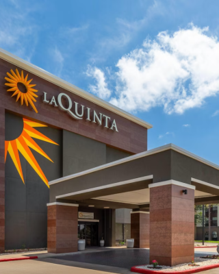 The image shows the exterior of a La Quinta hotel with a large logo on the building and a clear blue sky above.