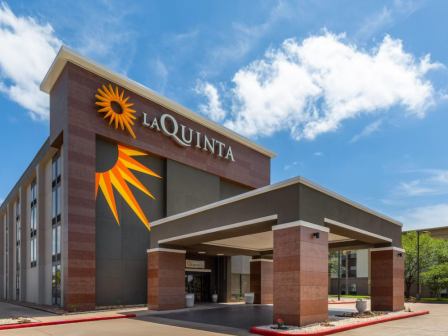 The image shows the exterior of a La Quinta hotel with a large logo on the building and a clear blue sky above.