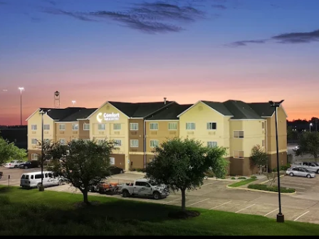 A hotel building with a "Comfort Suites" sign, surrounded by a parking lot and trees, at sunset.