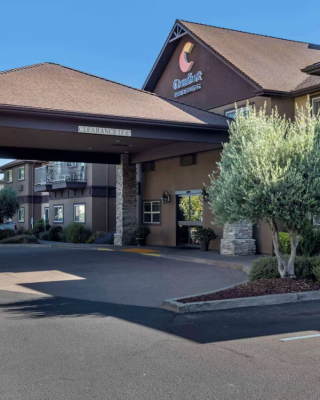 The image shows the exterior of a hotel with a covered entrance, landscaping, and parking spaces in front.