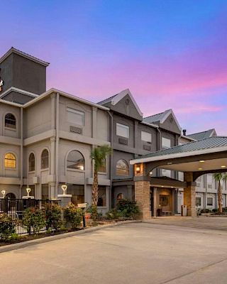 The image shows a hotel building with a parking area, surrounded by some landscaping, under a colorful sky during sunset or sunrise.