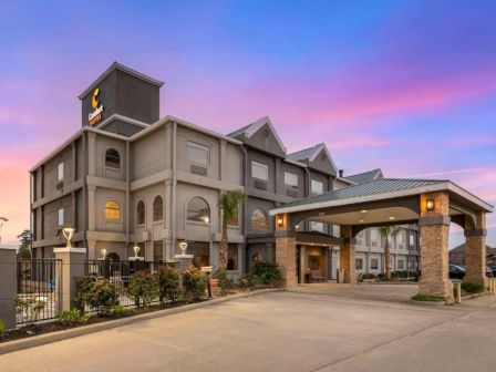 The image shows a hotel building with a parking area, surrounded by some landscaping, under a colorful sky during sunset or sunrise.