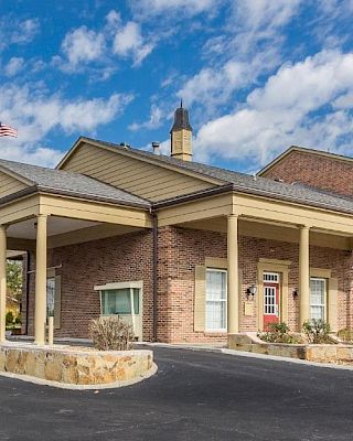 The image shows a brick building with a columned entrance and an American flag, set against a blue sky with clouds.