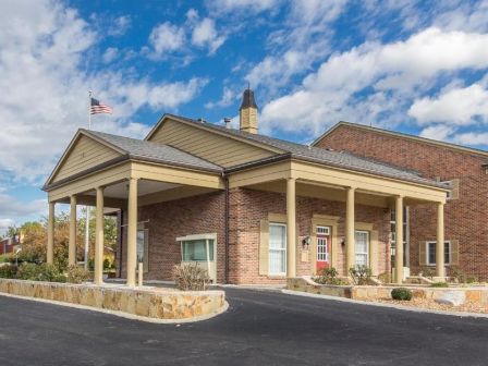 The image shows a brick building with a columned entrance and an American flag, set against a blue sky with clouds.