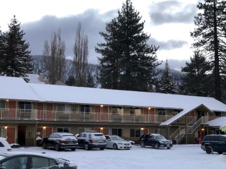 Snowy motel parking lot with a two-story beige building, pine trees, and several cars parked outside, winter scenery.
