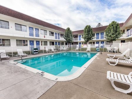 A rectangular communal pool surrounded by white lounge chairs, with a two-story beige apartment building and trees in the background.