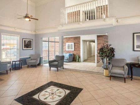 A spacious lobby with tiled floor, light blue walls, several chairs and small tables, brick accent walls, plants, and large windows. End.