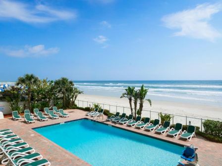 A sunny beachside pool area with a rectangular blue pool, numerous green lounge chairs around it, palm trees, and a sandy oceanfront beyond.