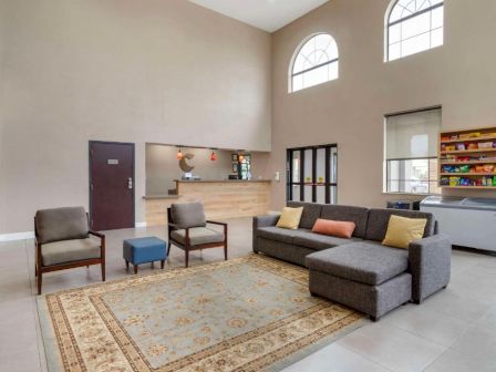 A bright, welcoming lobby with a gray sectional sofa, chairs, a coffee table, a reception desk, large windows, and a bookshelf by the wall.