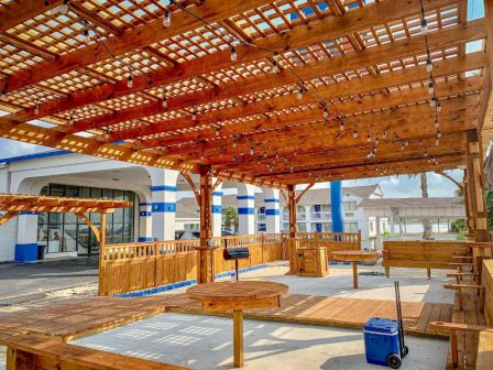 A wooden pergola-covered outdoor seating area with benches and tables, string lights, and a blue-and-white building in the background; sunny day.