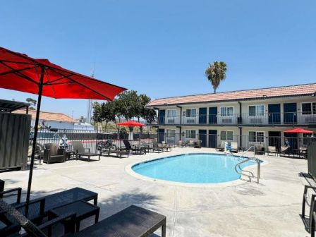 A sunny hotel courtyard with a small round pool, red umbrellas, lounge chairs, and a two-story building in the background.