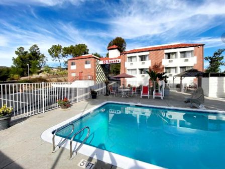 A sunny poolside at a motel/hotel with a rectangular blue pool, white railings, lounge chairs, and a two-story brick building in the background.