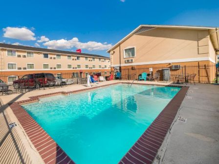 A rectangular outdoor hotel pool with turquoise water, surrounded by a brick coping, lounge chairs, tables, and a fenced patio area, under a clear blue sky.