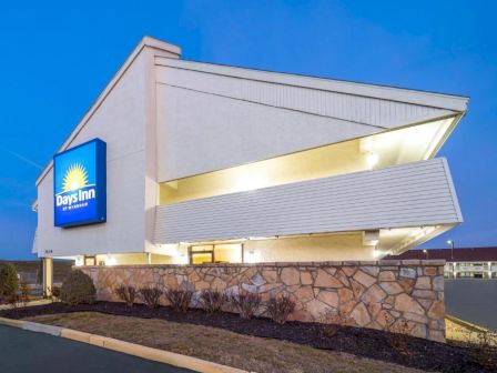 A modern Days Inn hotel with a angled white facade, stone lower wall, blue sign, and bright exterior lighting at dusk.