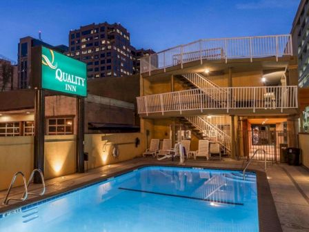 A nighttime scene of a Quality Inn with a lit outdoor pool, surrounding stairs, patio furniture, and a few tall buildings in the background, peaceful and inviting.