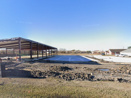 A construction site with a steel-framed shed, gravel ground, and blue tarp roofing under a clear sky.