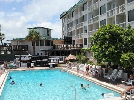 An outdoor hotel swimming pool with people swimming, surrounded by lounge chairs and apartment buildings, in sunny weather.
