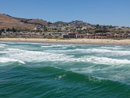An aerial view shows ocean waves rolling onto a sandy beach with a seaside town and hills in the background.