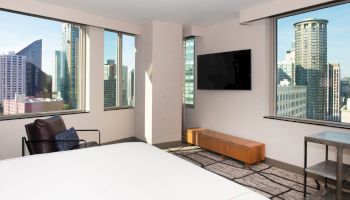 Modern hotel room with large windows, city view, armchair, wall-mounted TV, wooden cabinet, and a carpeted floor.