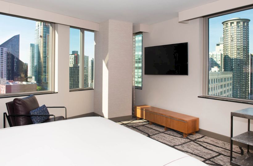 Modern hotel room with large windows, city view, armchair, wall-mounted TV, wooden cabinet, and a carpeted floor.
