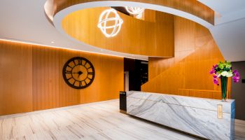 A modern lobby with a marble reception desk, wooden walls, decorative lighting, a large wall clock, and a vase with purple and white flowers.