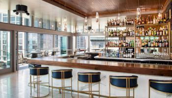 A modern bar with sleek counter seating, stocked with various liquors, and large windows offering city views.