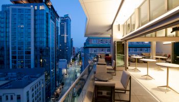 A modern cityscape at dusk with high-rise buildings and an elegant balcony featuring outdoor seating and bright interior lighting.
