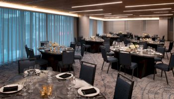 A beautifully set dining room with round tables, black chairs, plates, glasses, and floral centerpieces, ready for an event or gathering.