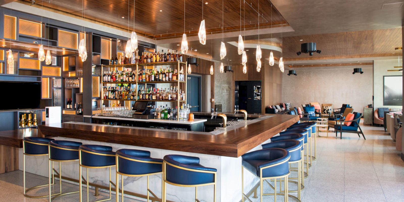 A modern bar with a wooden counter, blue chairs, pendant lights, and a fully stocked liquor shelf. Seating area visible in the background.