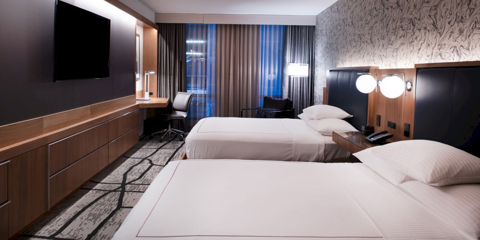 A modern hotel room with two beds, a TV, desk, and decorative wallpaper. The room is well-lit with lamps and features a carpeted floor.