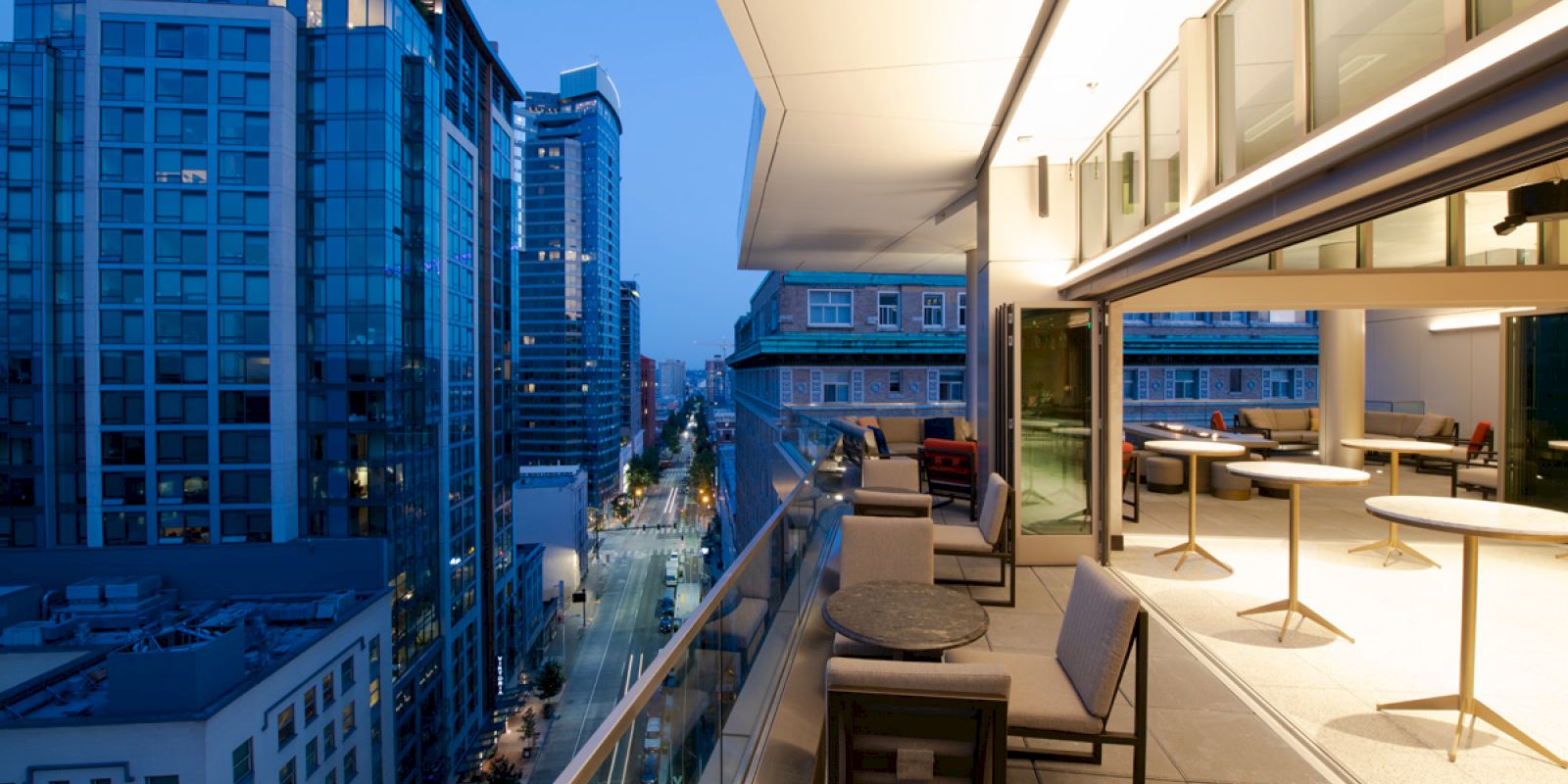 Rooftop terrace with seating overlooks a city street and modern buildings in the evening.