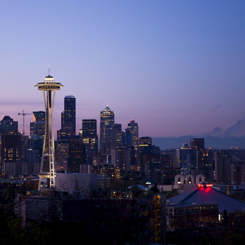 City skyline at dusk with the Space Needle and Mount Rainier in the background, featuring lit buildings and a clear sky.