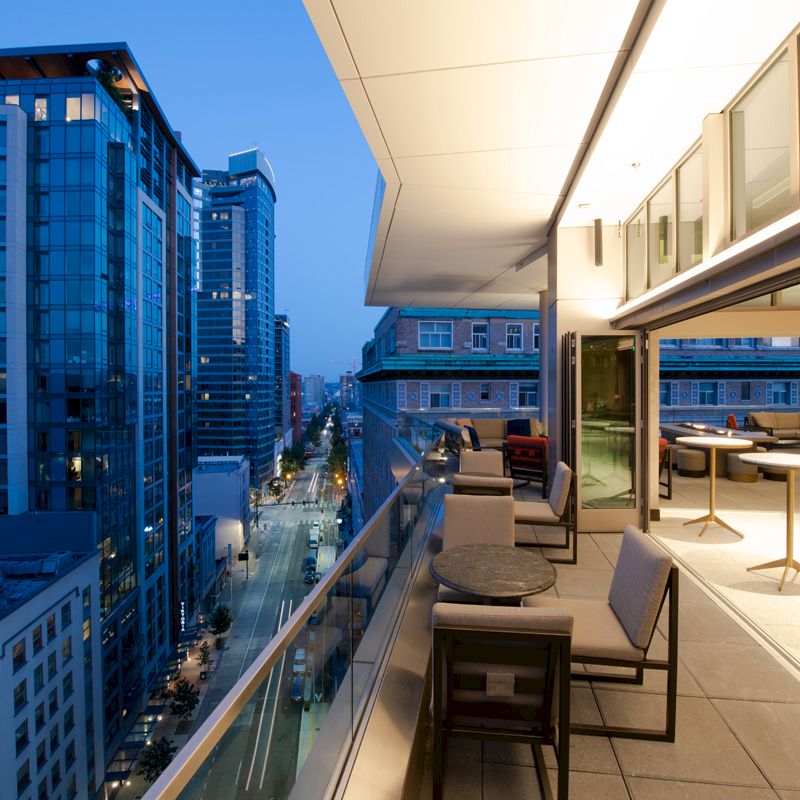 A modern rooftop patio with tables and chairs overlooks a city street lined with tall buildings during early evening.