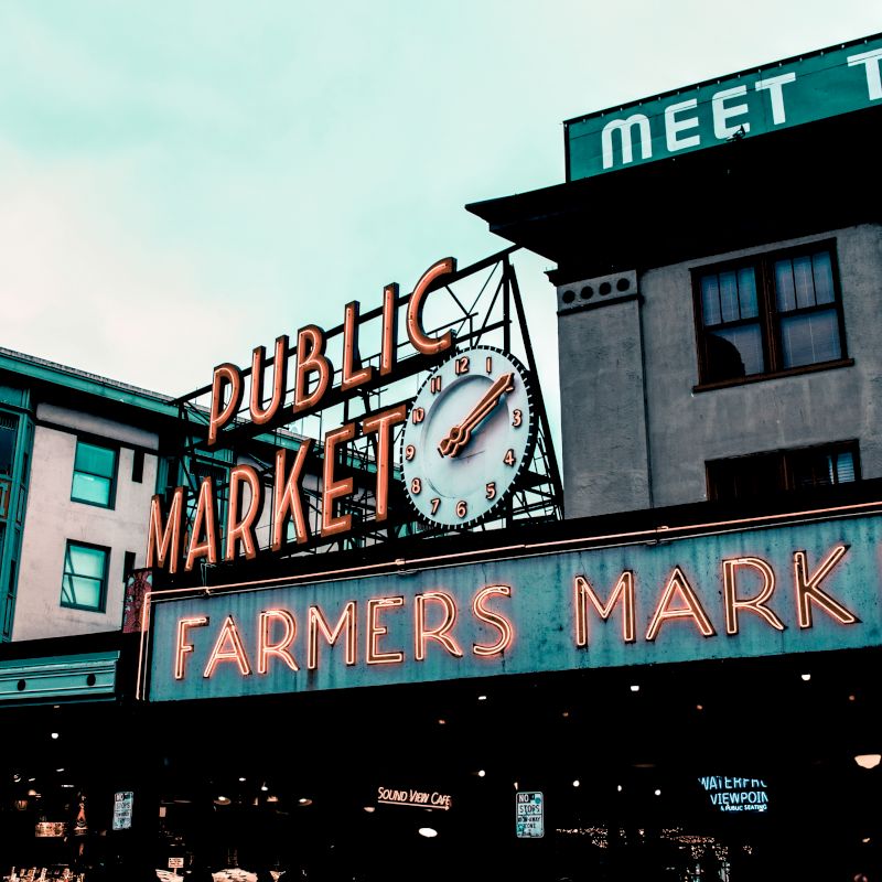 The image shows a neon sign for a public farmers market, attached to a building, with a clock in the center and cloudy sky above.